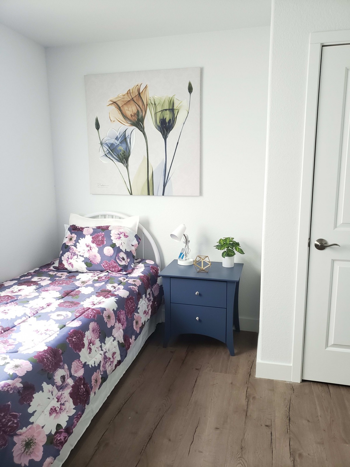 A minimalist bedroom with a single bed featuring floral bedding, a nightstand with a lamp and plant, and vibrant artwork on the wall.