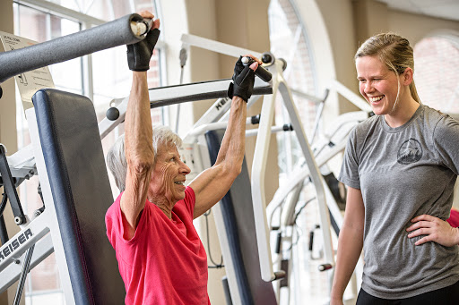 An older adult engages in strength training with the assistance of a personal trainer at the gym.