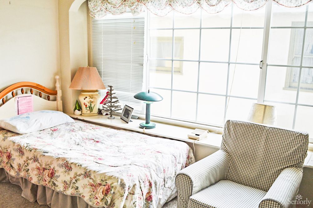 A cozy bedroom with a bed, lamp, and armchair next to a large window.