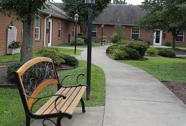 A peaceful garden setting with a bench beside a concrete pathway leading to brick buildings.