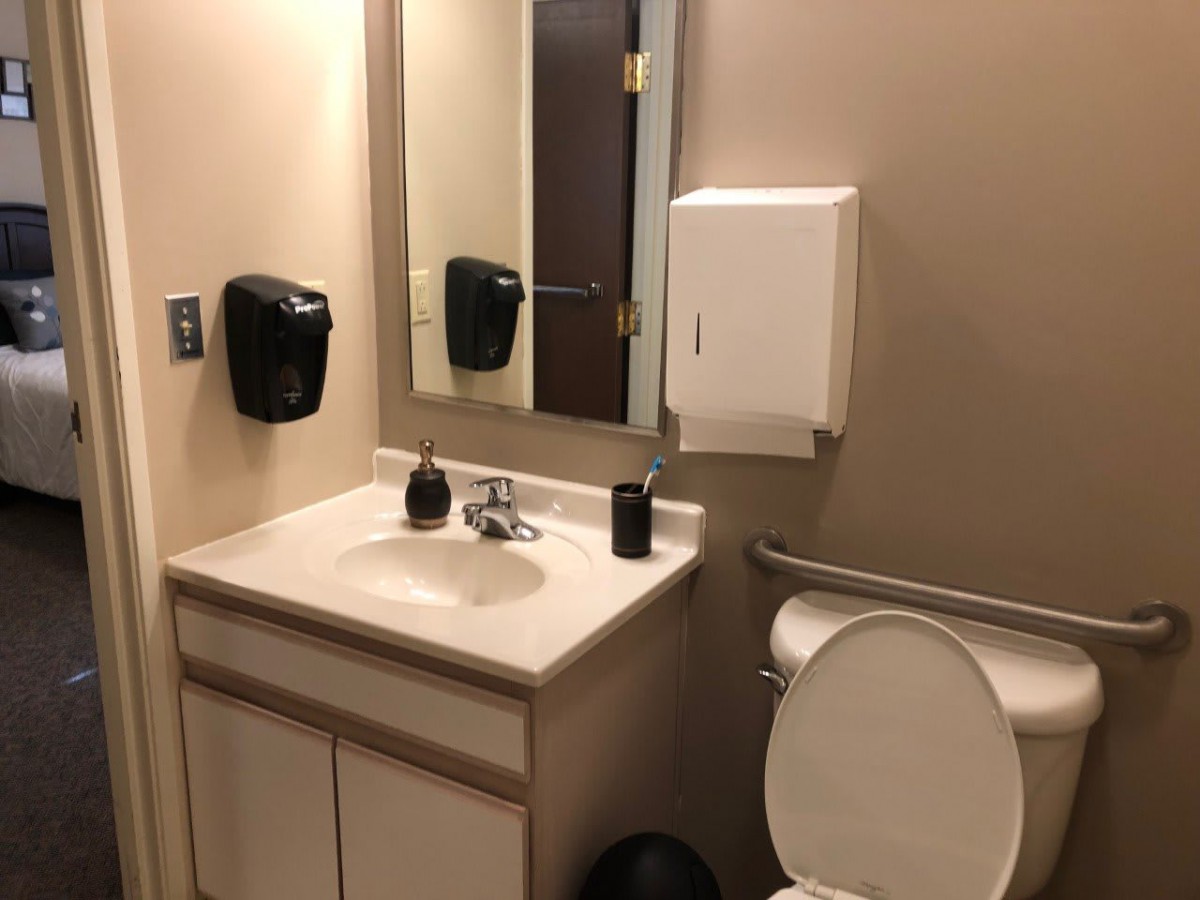A clean and accessible bathroom with a sink, mirror, toilet, and dispensers.