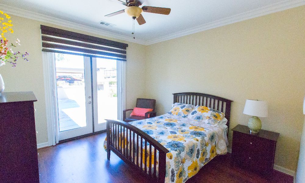A cozy bedroom with a wooden bed frame, floral bedspread, ceiling fan, and a window with curtains.