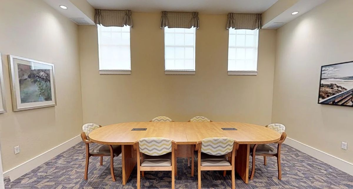 A conference room with a large wooden table and several chairs, positioned below three windows.