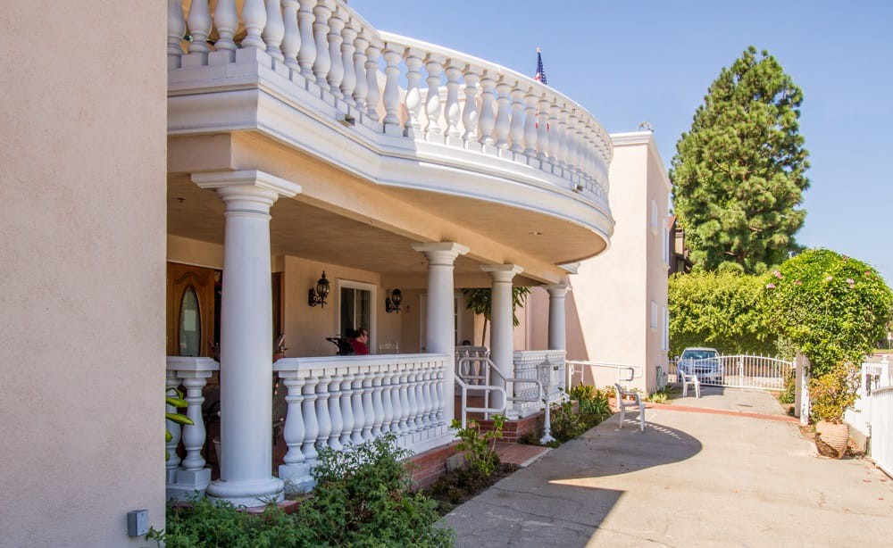 A large house featuring a grand balcony with ornate columns and a garden-lined driveway.