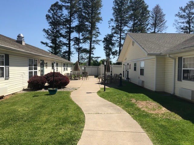 A sunny day with a pathway between two buildings, surrounded by grass and tall trees.
