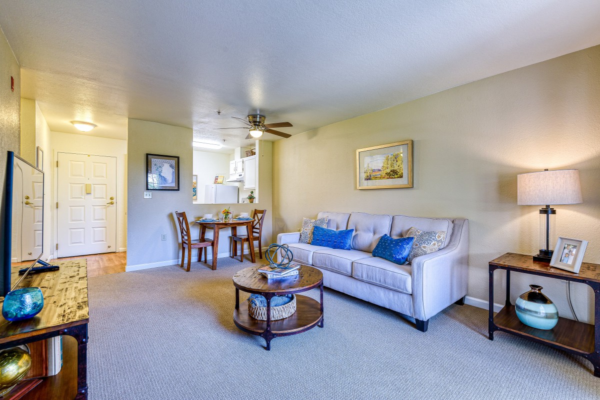 A cozy living room with a sofa, coffee table, and dining area, featuring a ceiling fan and wall art.
