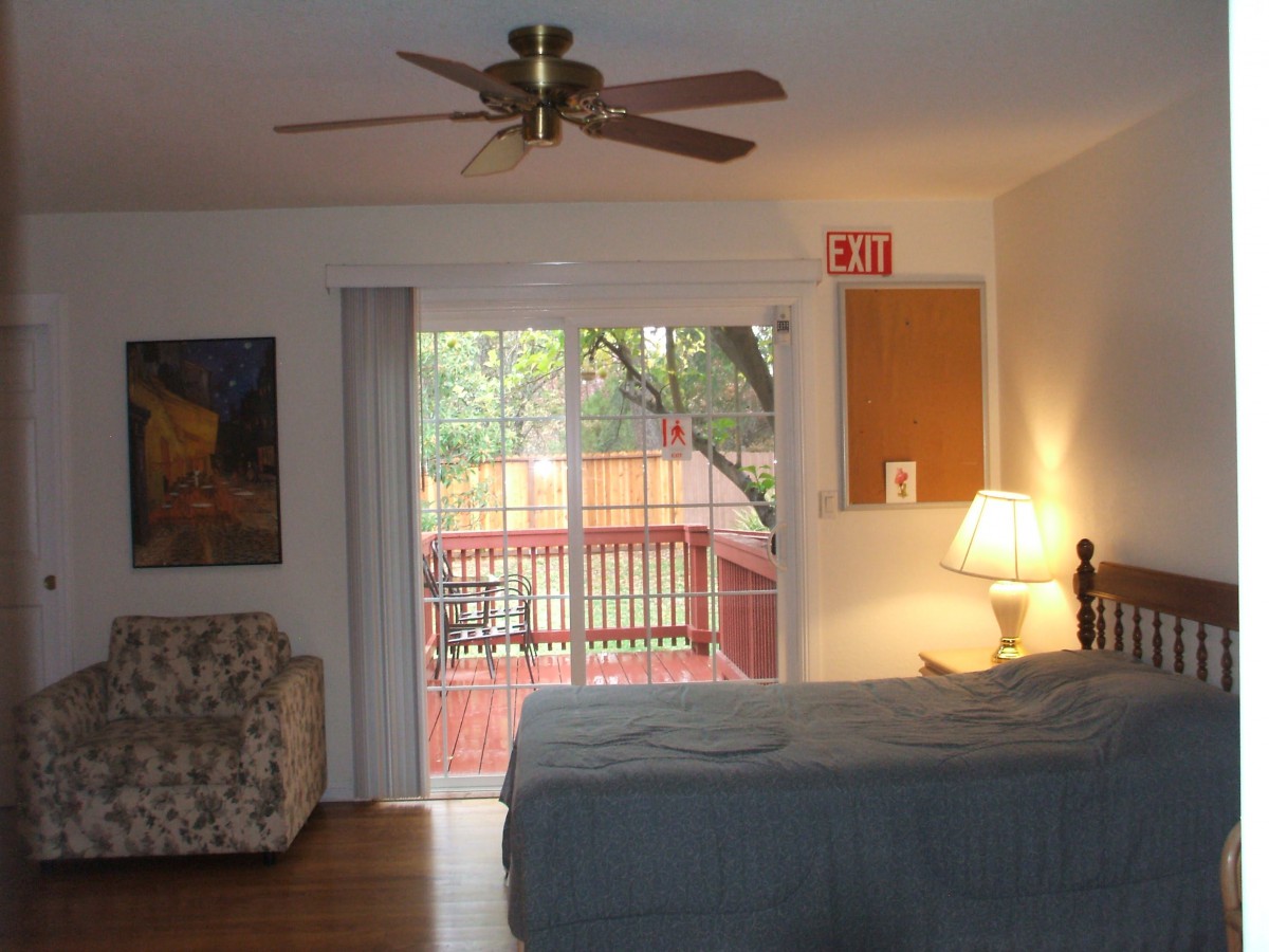 A cozy bedroom with a ceiling fan, bed, and lamp, featuring a sliding glass door opening onto a deck with natural light.