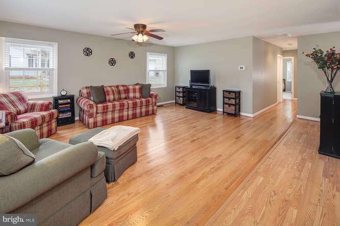 A spacious living room with hardwood flooring, featuring a plaid sofa, armchair, ceiling fan, and a television set.