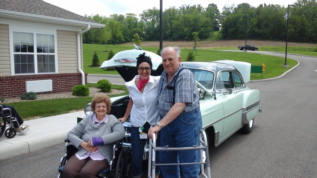 Three people posing outside with a classic car, including an elderly couple using mobility aids.