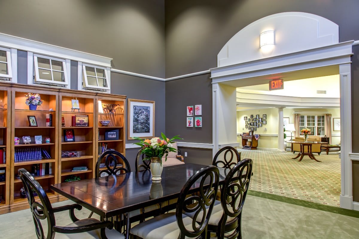 A well-lit dining area featuring a dark wooden table, chairs with ornate backs, a vase of flowers, and decorative elements including a bookshelf and framed artwork.