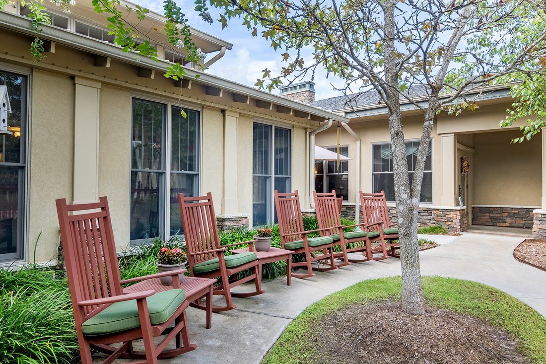 A row of wooden rocking chairs with green cushions outside a building surrounded by a garden.