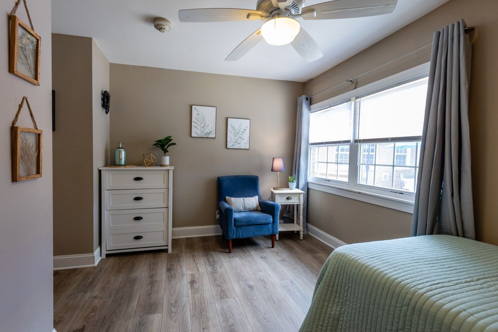 A cozy bedroom with a blue chair, dresser, and large window letting in natural light.