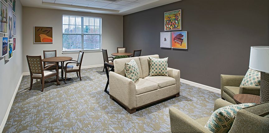 A cozy living room featuring a beige sofa with patterned cushions, wall art, and ample natural light from a large window.