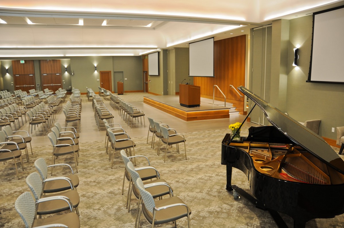 An empty conference room with rows of chairs, a stage with a podium, projector screens, and a grand piano.