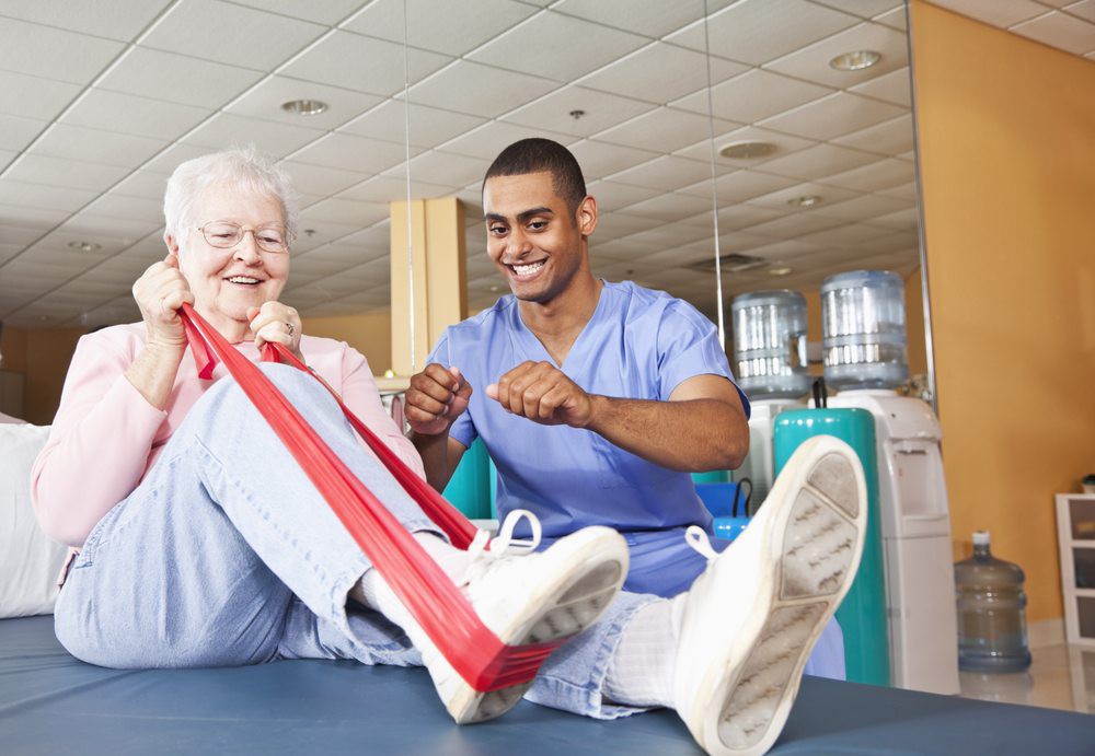 An elderly woman is assisted by a healthcare professional during a physical therapy session using an exercise band.
