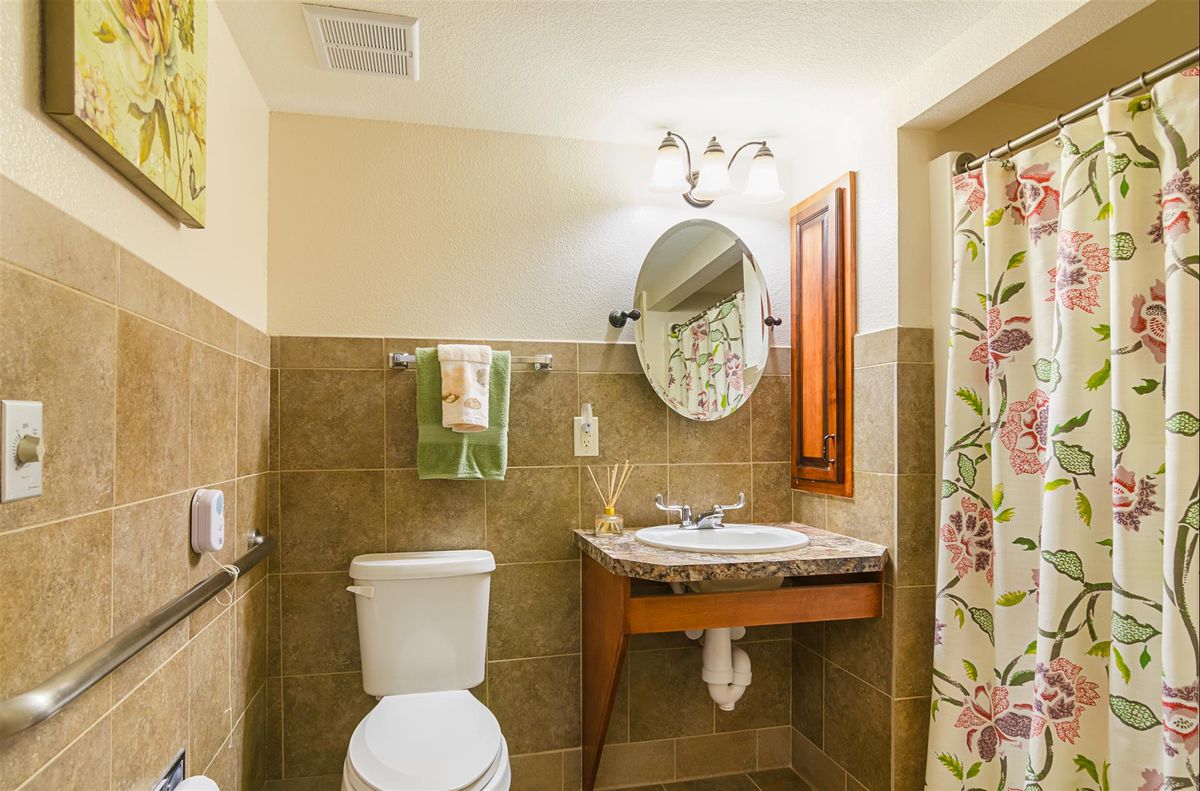 A well-lit bathroom featuring a toilet, sink with an oval mirror, and a floral shower curtain.