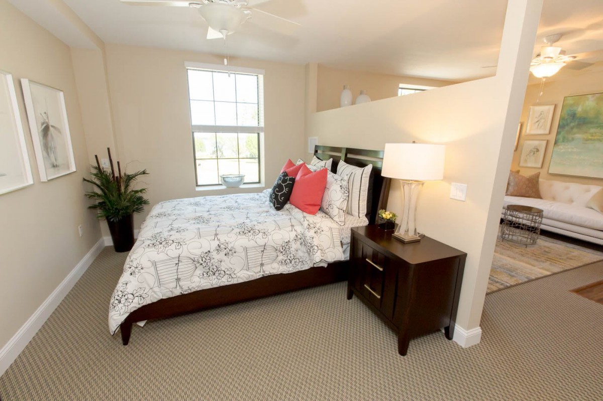 A modern bedroom featuring a neatly made bed with floral bedding, flanked by a nightstand and lamp, complemented by a large window and decorative plants.