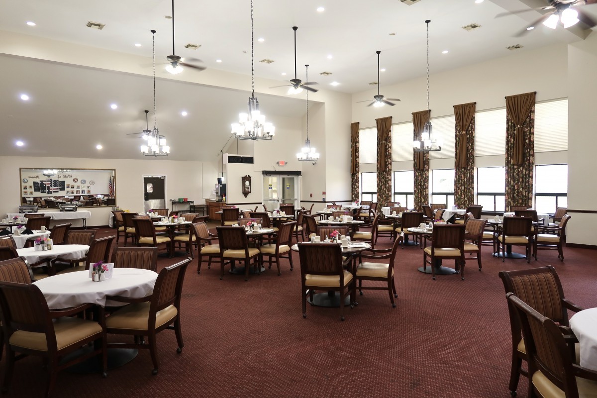 A spacious dining hall with elegant chandeliers, carpeted flooring, and neatly arranged tables and chairs.