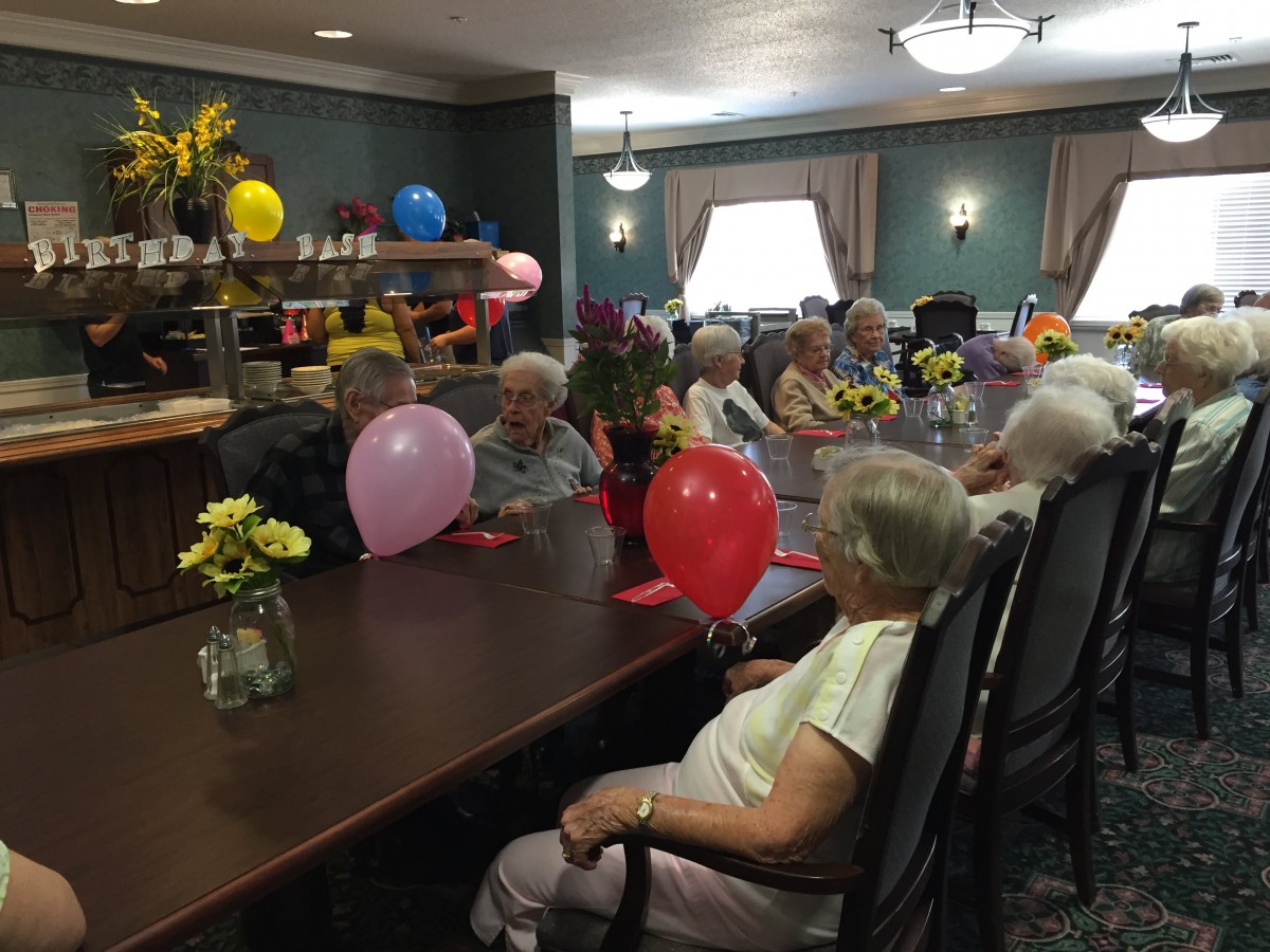 A group of seniors enjoying a celebration with balloons and flowers in an indoor setting.