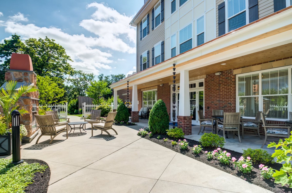 A sunny outdoor patio area with chairs and tables, adjacent to a multi-story building surrounded by landscaped greenery.