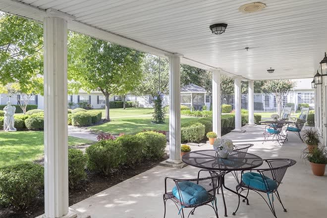 A covered patio with outdoor furniture overlooks a well-manicured garden featuring trees and shrubs.