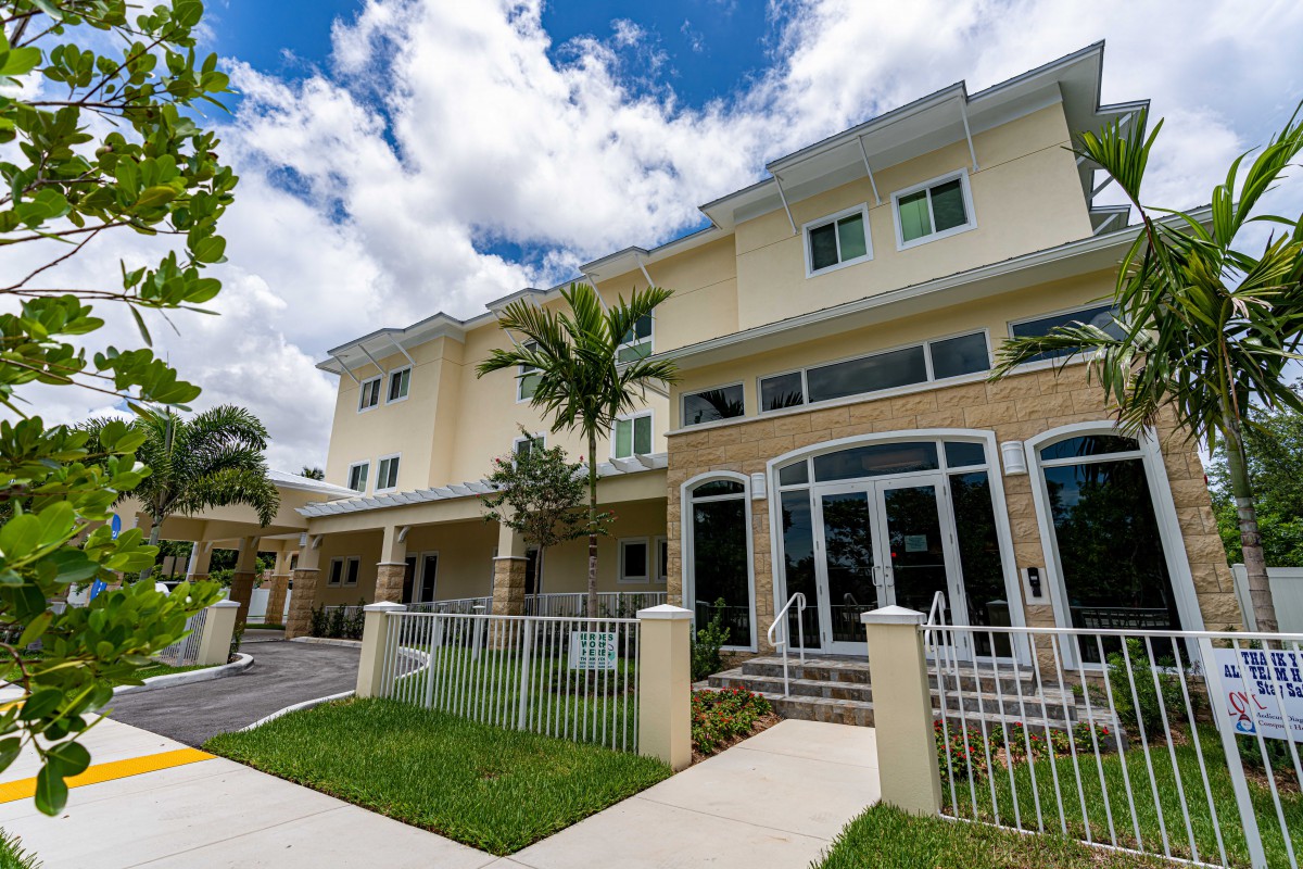 A modern residential building with large windows and a small front yard, set against a partly cloudy sky.