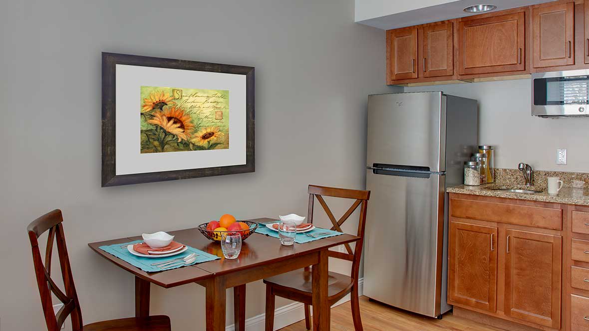 A cozy kitchen corner featuring a small dining table set with place settings, a bowl of fruit, and a sunflower painting on the wall.