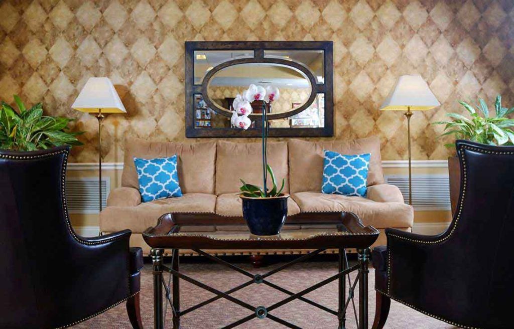 A cozy living room interior with a central coffee table adorned with an orchid plant, a beige sofa, and blue patterned throw pillows, accented by two floor lamps and a decorative mirror on the wall.