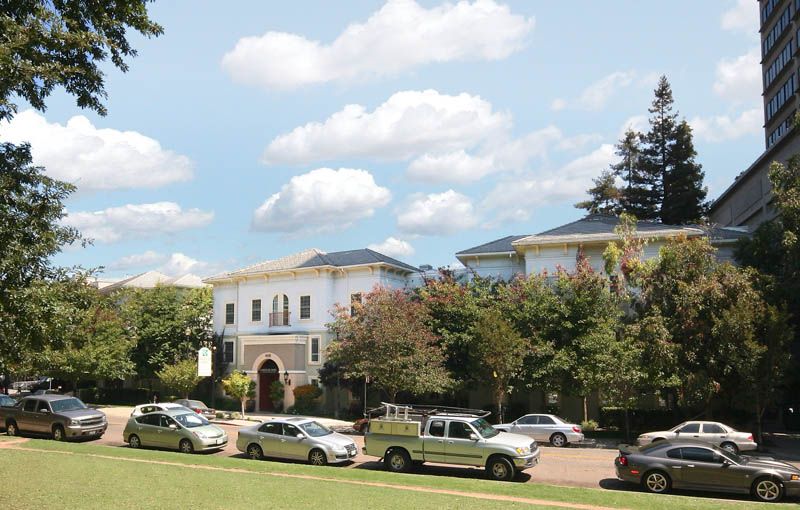 A view of a stately building with a row of cars parked in front, surrounded by trees against a backdrop of blue sky and clouds.