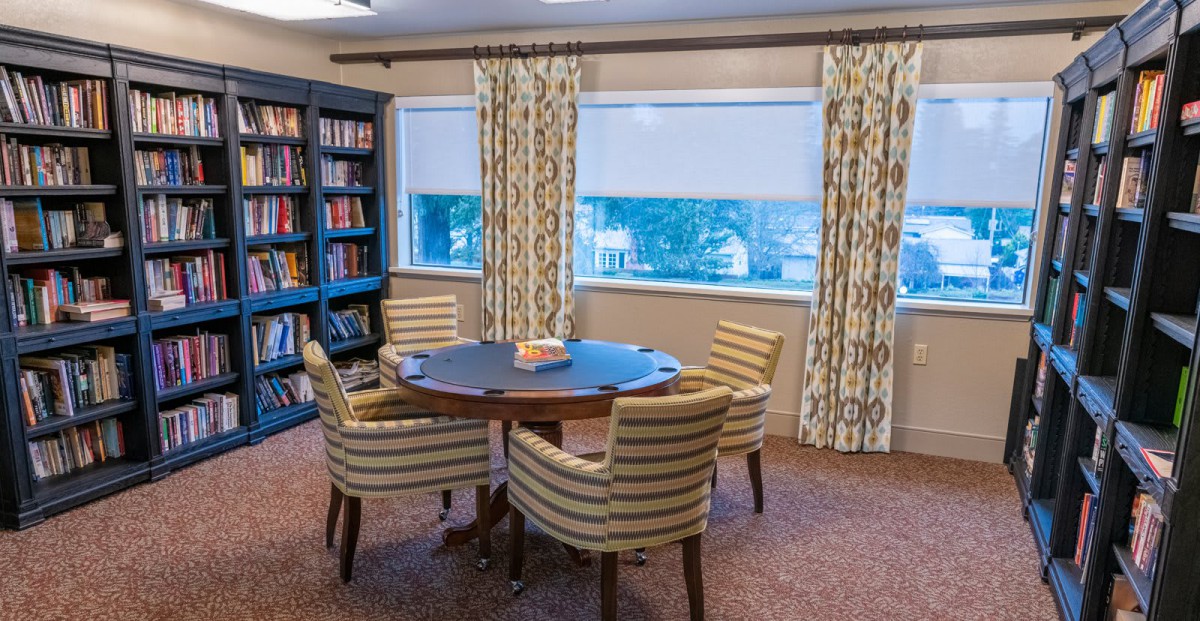A cozy reading room with a round table and striped chairs, surrounded by bookshelves and windows.