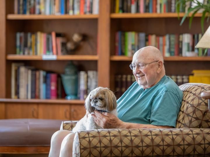 An elderly person sitting in a chair with a dog on their lap, surrounded by bookshelves.