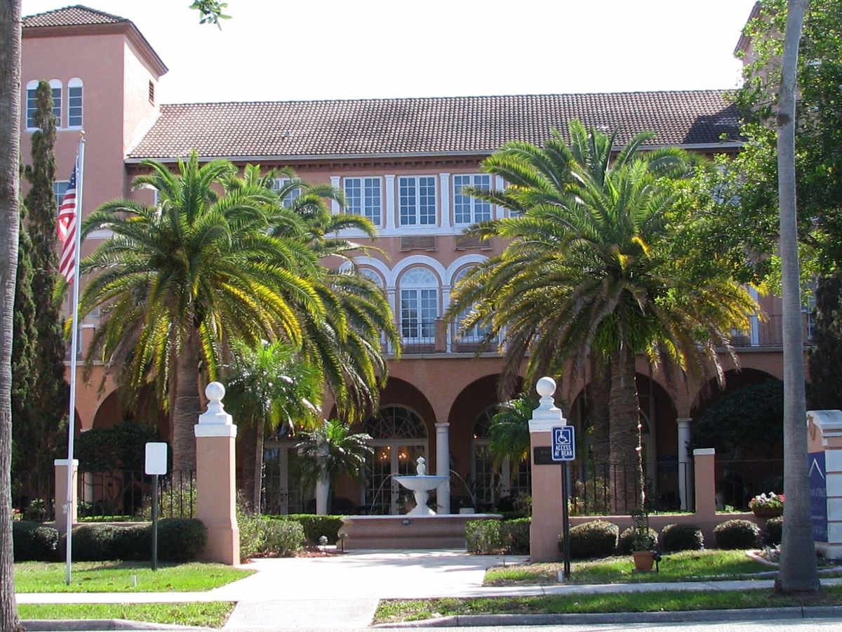A building with arched windows and palm trees at the entrance, flanked by statues.