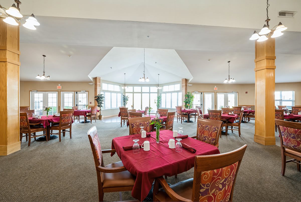 A spacious dining room with tables and chairs arranged neatly, featuring red tablecloths and large windows providing natural light.