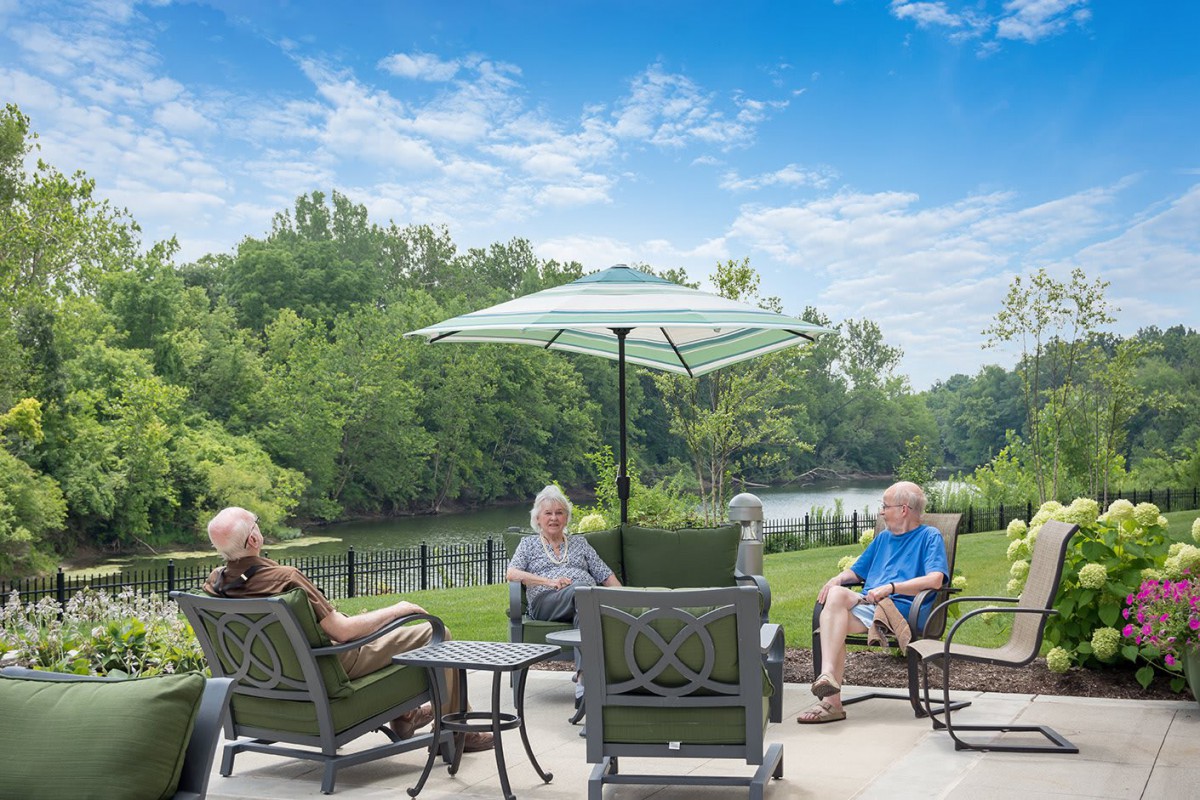 A group of people enjoying a relaxing afternoon on a patio by the river, surrounded by trees.