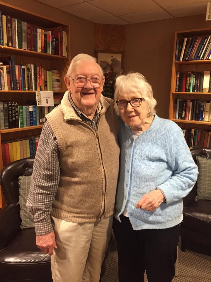 An elderly couple smiling together in front of a bookshelf.