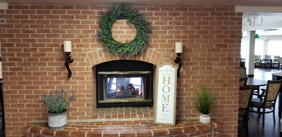 A cozy brick fireplace decorated with a wreath, home sign, candle, and indoor plant.
