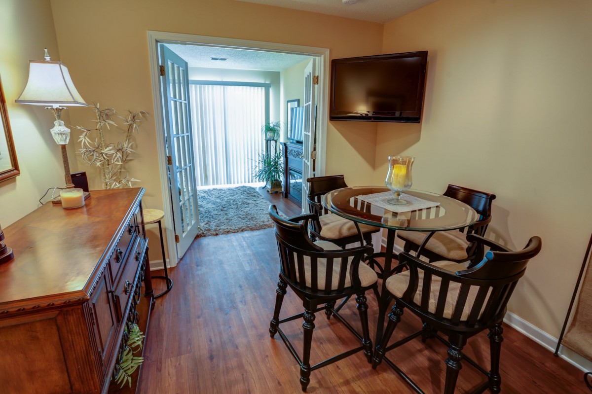A cozy dining area with a round table and chairs, a mounted TV, and glass doors leading to another room with indoor plants.