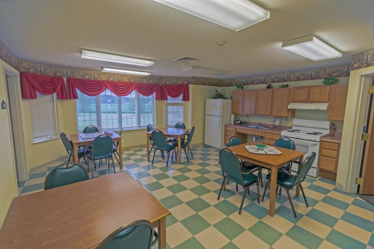 A communal kitchen and dining area with multiple tables and chairs, featuring large windows and a checkered floor.
