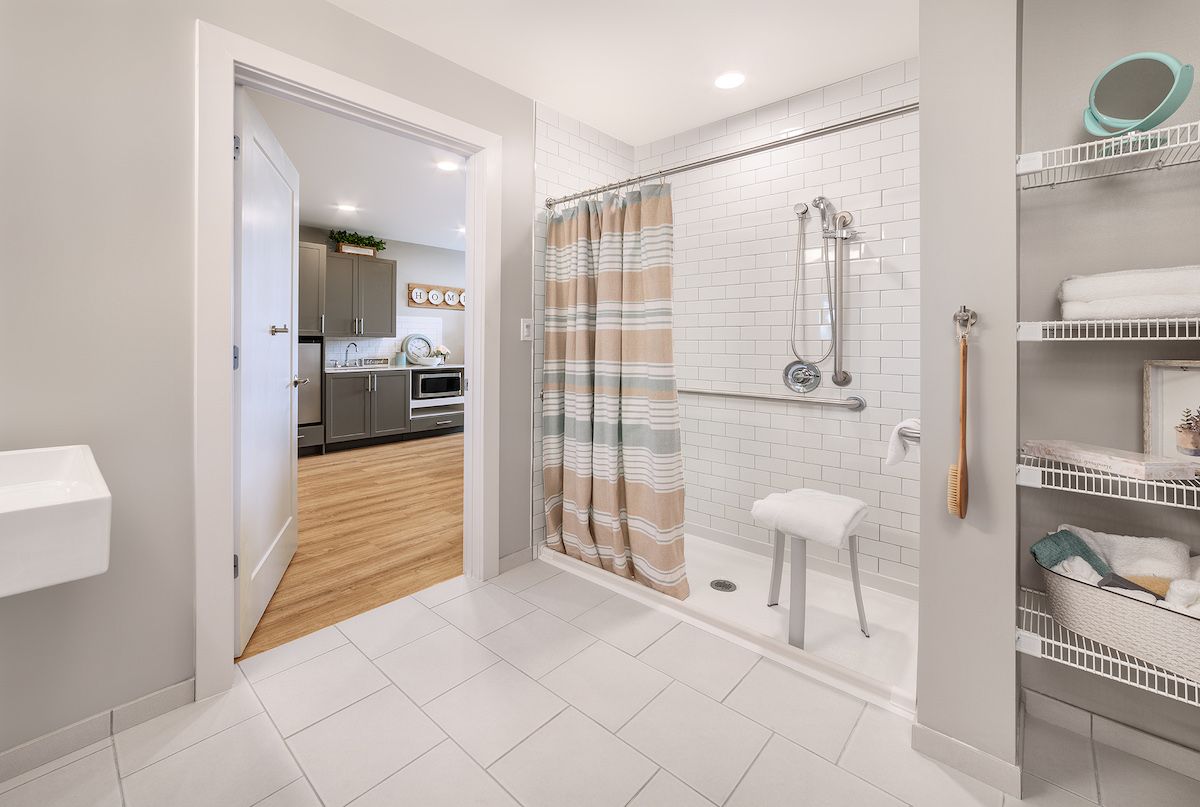 A modern bathroom featuring an accessible shower with a striped curtain and a view into the kitchen.