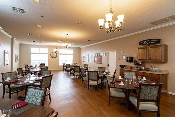 A well-lit dining room with neatly arranged tables and chairs, featuring elegant chandeliers and wooden flooring.