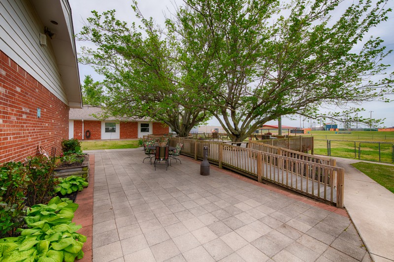 A spacious backyard with a paved patio area featuring outdoor seating and a large tree, surrounded by a fence.