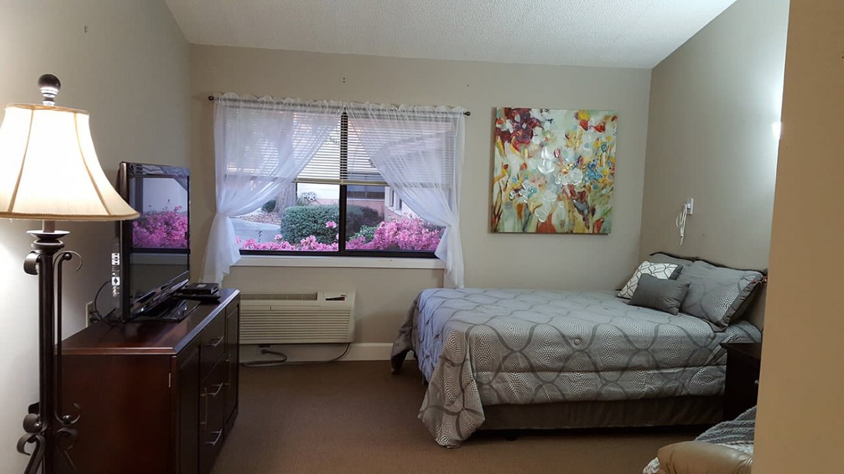 A cozy bedroom with a large window, featuring a bed, dresser, and colorful artwork on the wall.