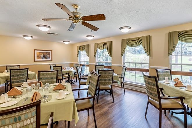 A cozy dining room with neatly arranged tables and chairs, featuring large windows with draped curtains and a ceiling fan.