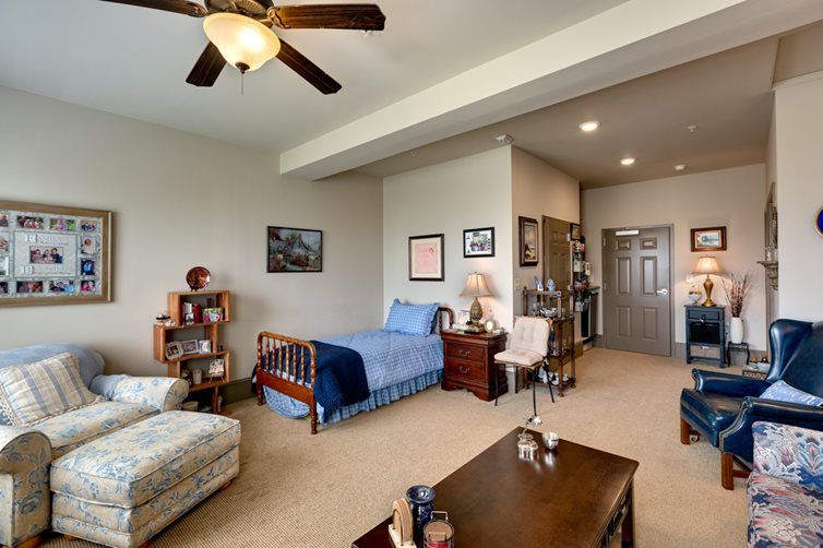 A cozy bedroom featuring a single bed, bookshelf, and armchair with a ceiling fan overhead.