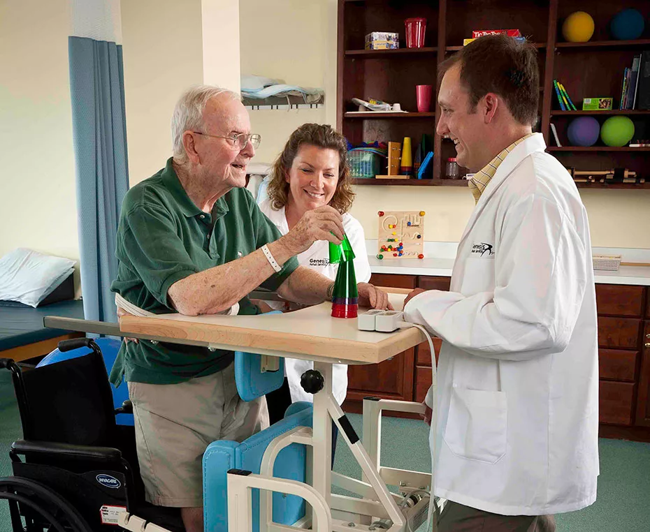 An elderly man receives assistance from a medical professional and caregiver during a physical therapy session.