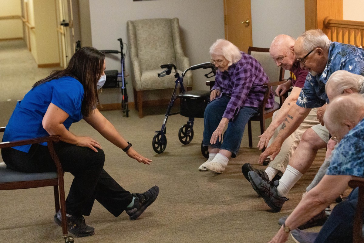A group of seniors participating in a stretching exercise led by an instructor in a care facility.