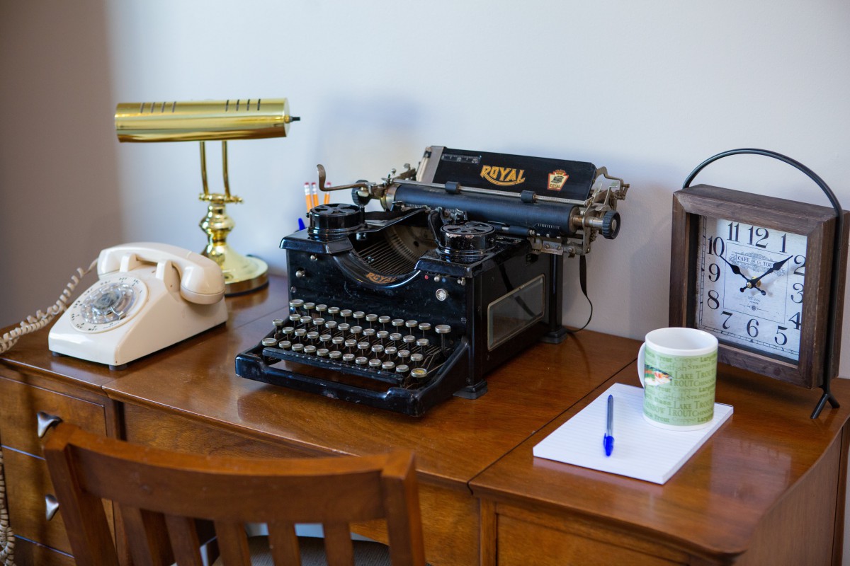 A vintage typewriter sits on a wooden desk alongside a lamp, notepad, pen, and mug.