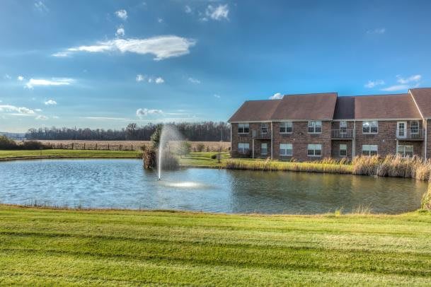 A serene pond with a fountain alongside a brick building under a clear blue sky.