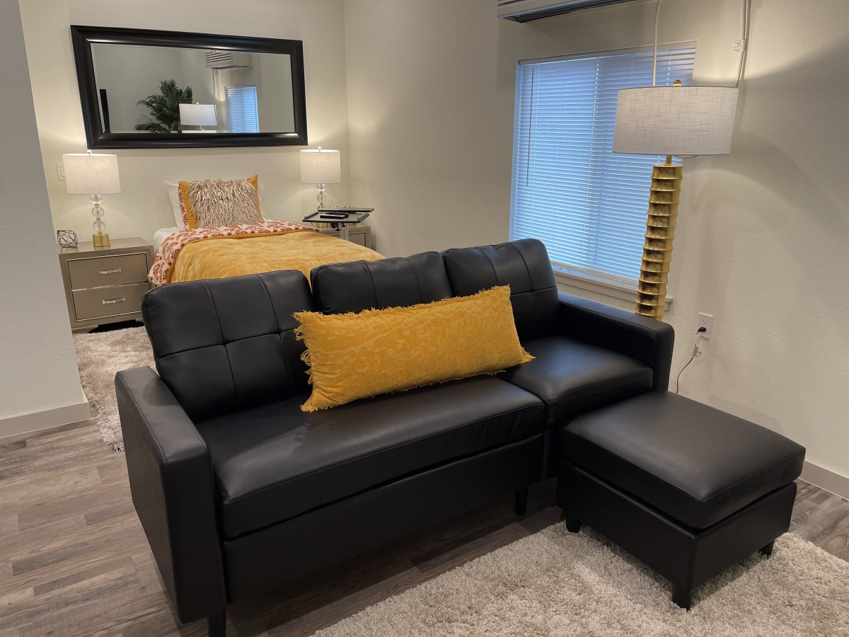 A cozy living room with a black sofa and ottoman, complemented by a yellow pillow, near a bed with warm lighting.