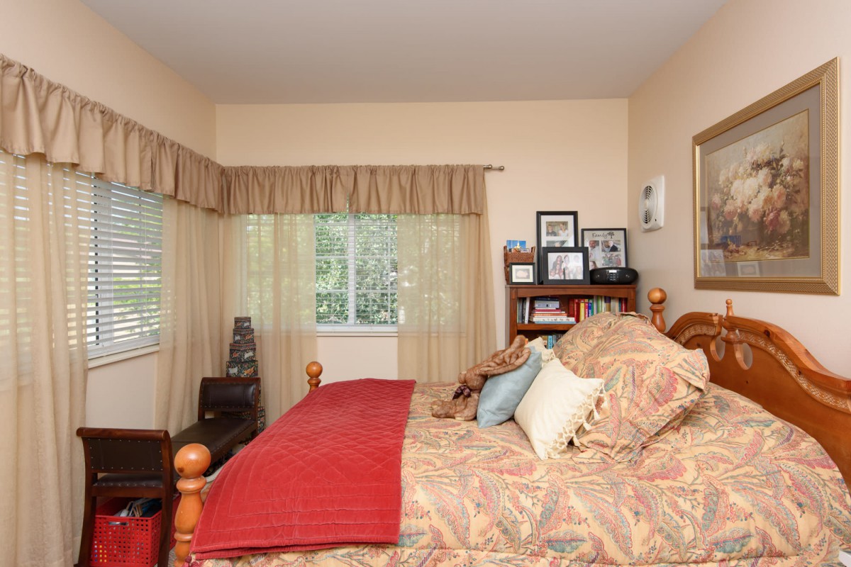 A cozy bedroom with a wooden bed, patterned bedspread, red blanket, and decorative pillows, featuring shelves with framed photos by the window.